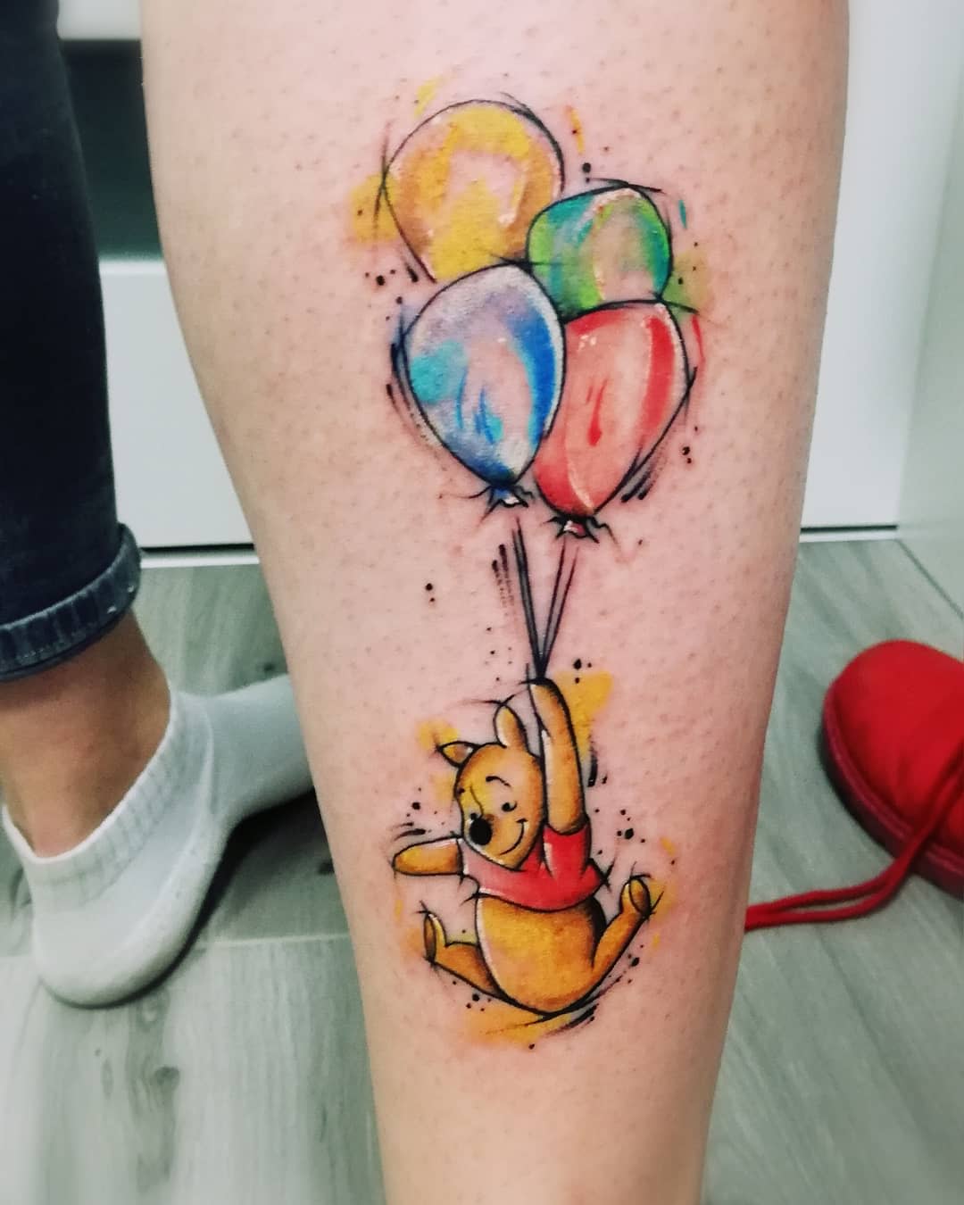 Watercolor tattoo of Winnie the Pooh holding colorful balloons on a calf